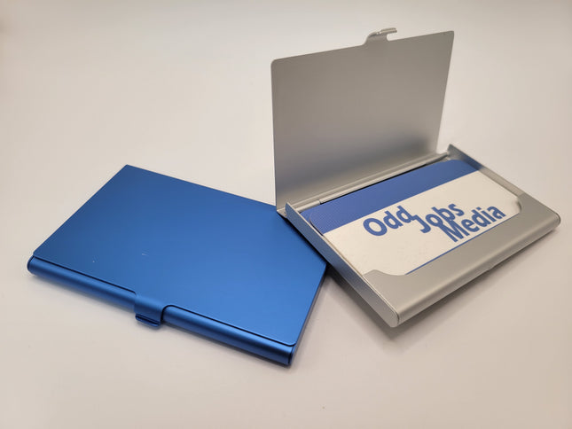 A metallic blue aluminum business card holder shown closed next to an open silver version containing a branded card that reads 'Odd Jobs Media'.