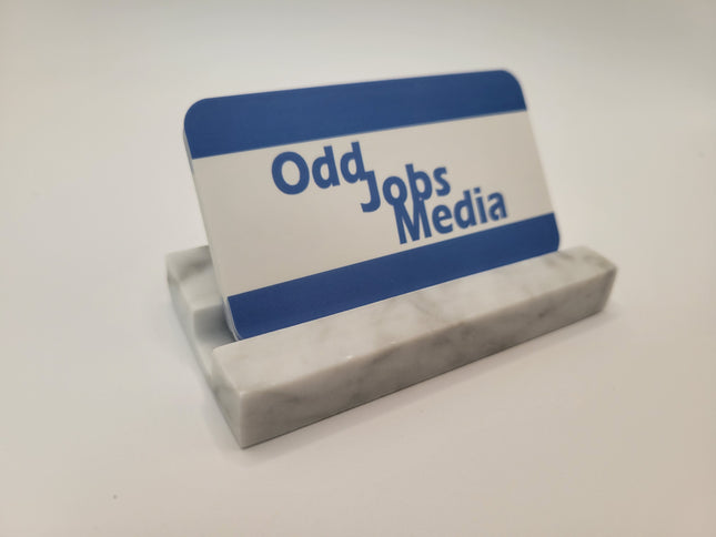 A small rectangular business card on a marble stand with blue and white horizontal bands and the text 'Odd Jobs Media' printed on it, photographed on a plain light background.