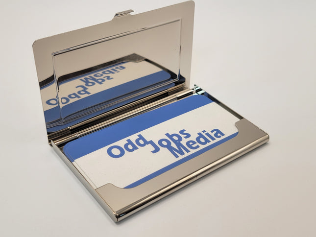 A polished silver metal business card holder, shown open to display a custom printed card inside. The card features a white background with a blue curved stripe and the text 'Odd Jobs Media' in blue.
