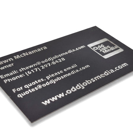 [photo] A close-up of a black business card with white text showing contact details and the Odd Jobs Media logo, photographed on a pure white background.