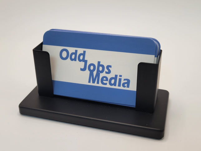 A black steel business card holder displaying blue and white business cards with the text 'Odd Jobs Media' on a light gray background.